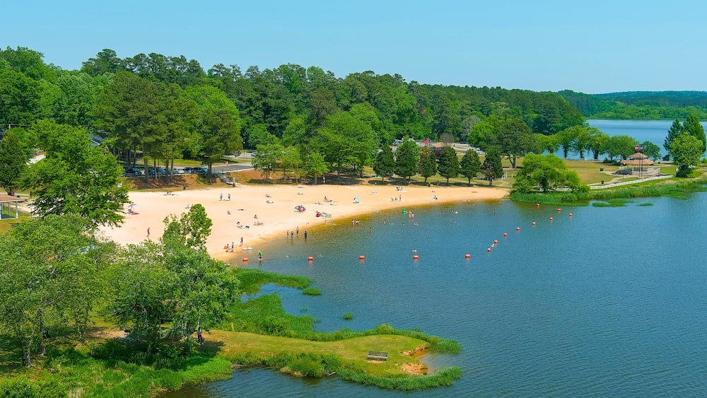 an aerial shot of people relaxing on the beach at Lake Acworth with silky brown sand and blue lake water surrounded by lush green trees and grass with blue sky at Cauble Park in in Acworth Georgia USA
