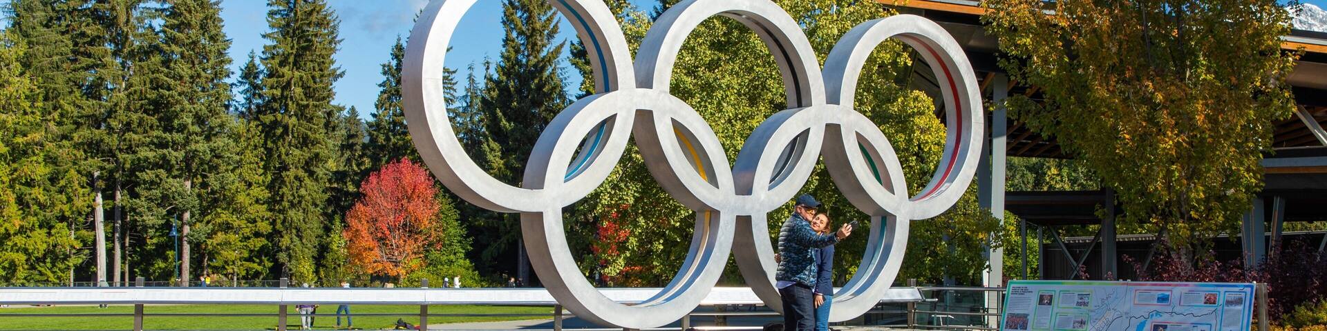 A large Olympic rings sculpture in a park with trees and a clear sky.