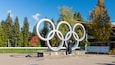 Une grande sculpture représentant les anneaux olympiques, située dans un parc avec des arbres et sous un ciel dégagé.