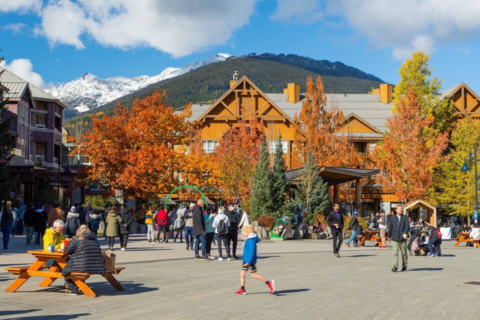 Una plaza animada con edificios de madera, árboles otoñales y montañas al fondo.
