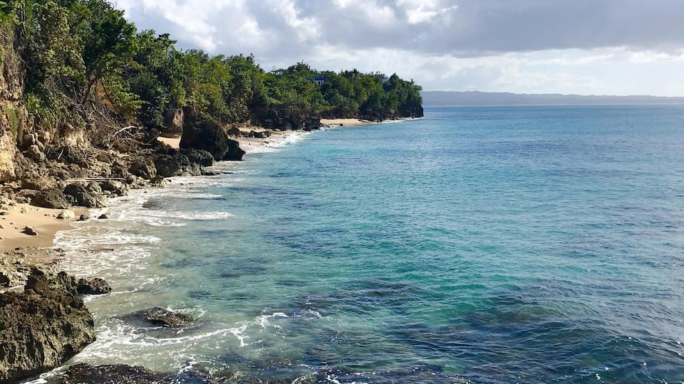 A coastal landscape with clear blue waters, rocky shores, and dense green vegetation.