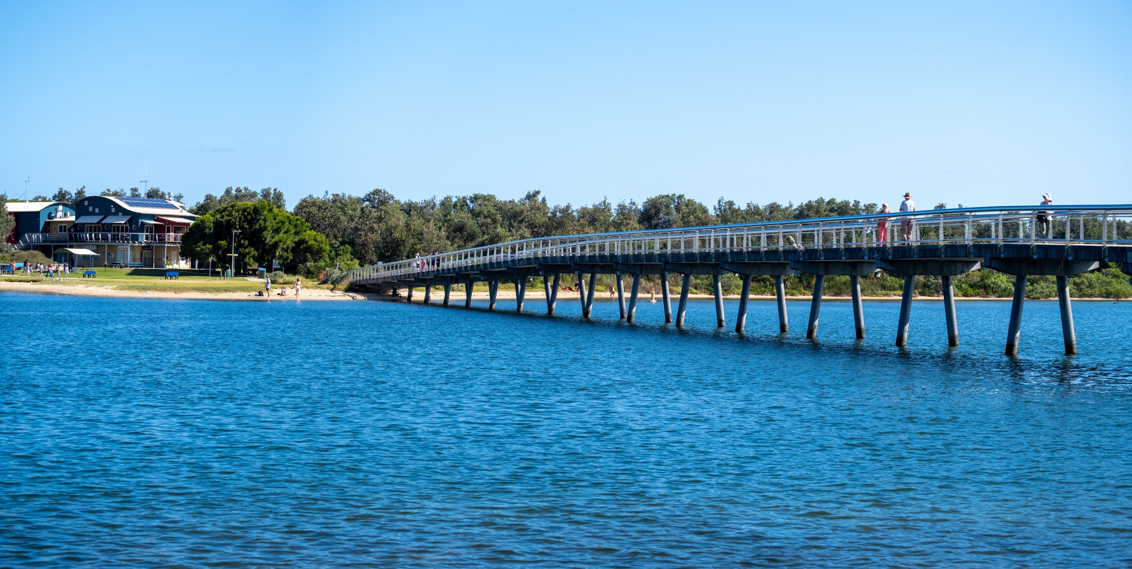 Panoramic view of the Cunninghame Arm Footbridge stretches across the calm blue water of Lake Entrance. People stroll along the bridge, enjoying the scenic views and the summer holiday