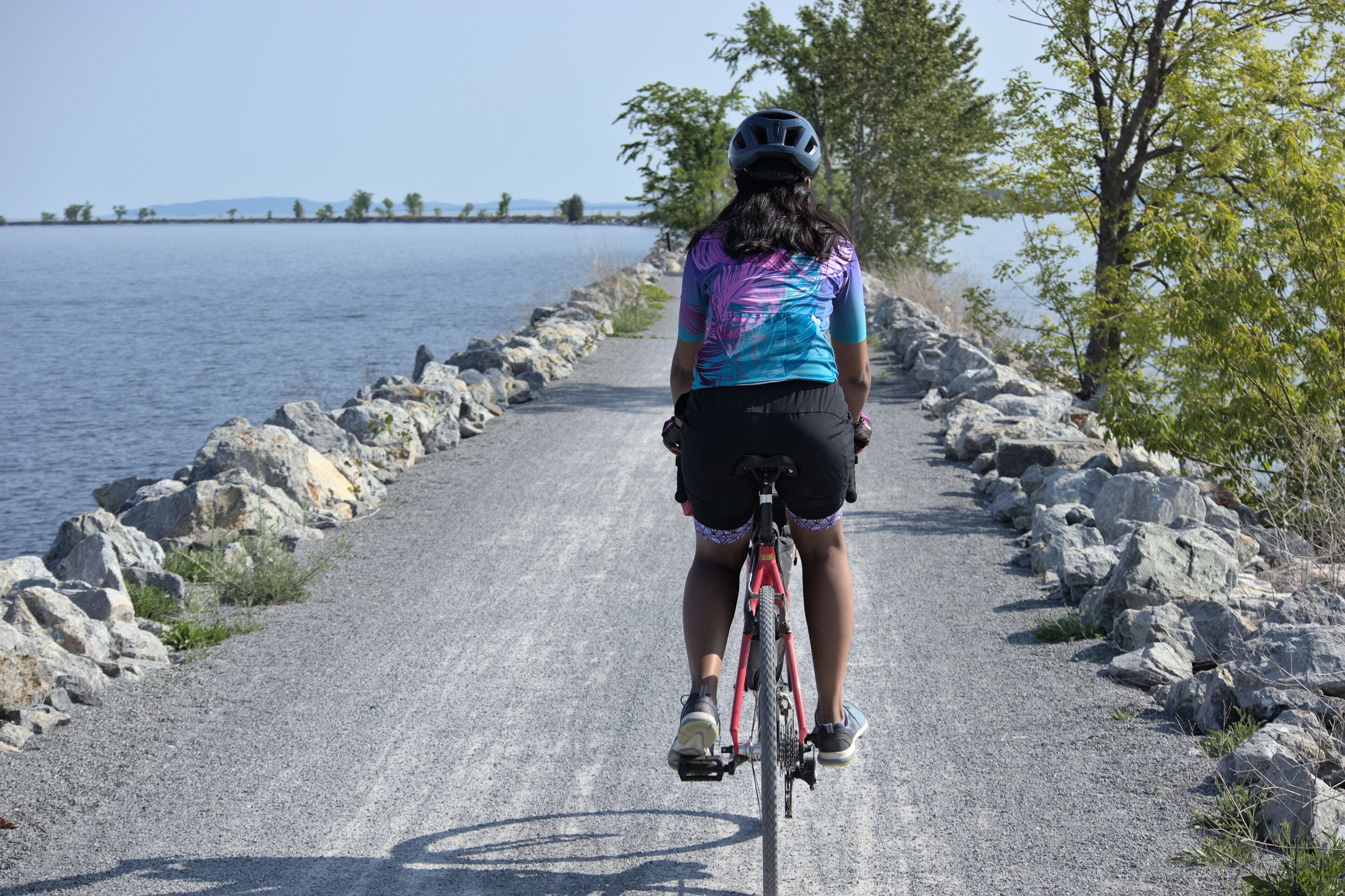woman riding bike on gravel trail (young south asian, indian rider on bicycle trail) burlington vermont colchester causeway path (brown skin, athletic clothes, cycling jersey, biking shorts, helmet)
