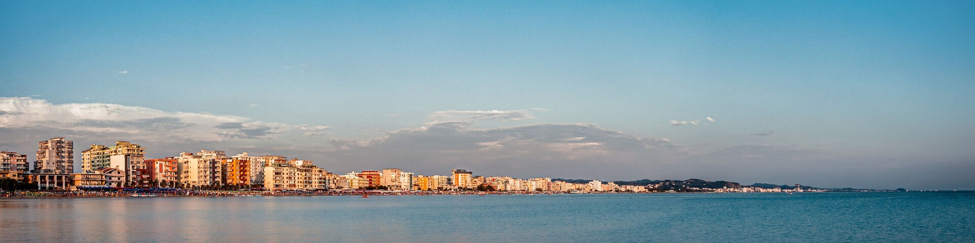 A coastal cityscape with a clear sky, calm sea, and a variety of buildings along the waterfront.