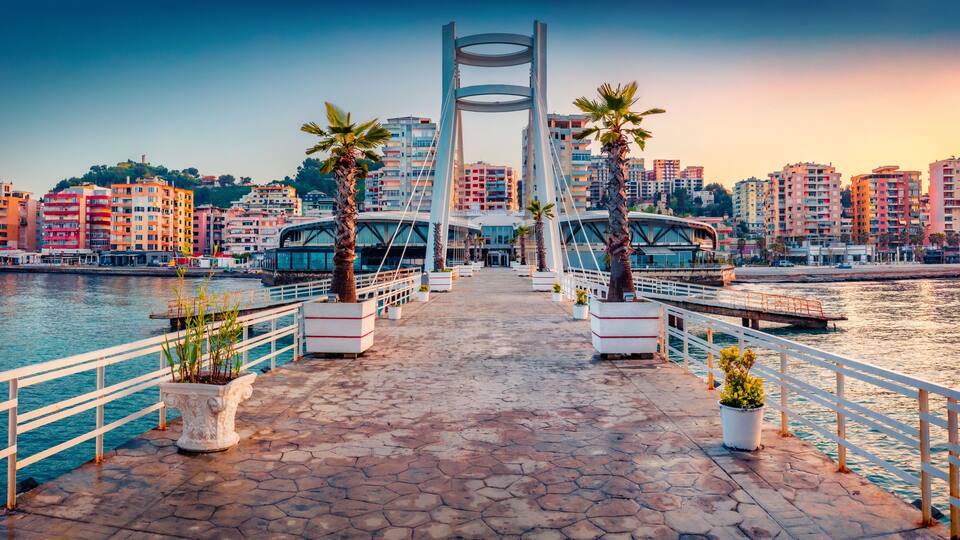A waterfront promenade with a modern bridge, palm trees, and residential buildings.