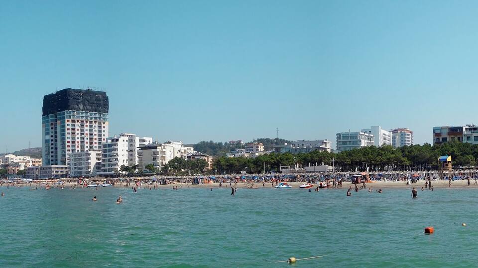 A coastal cityscape with a beach, boats, and a prominent skyscraper.