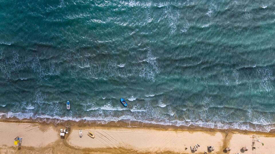 An aerial view of a beach with waves crashing onto the shore, a few boats, and beach umbrellas.