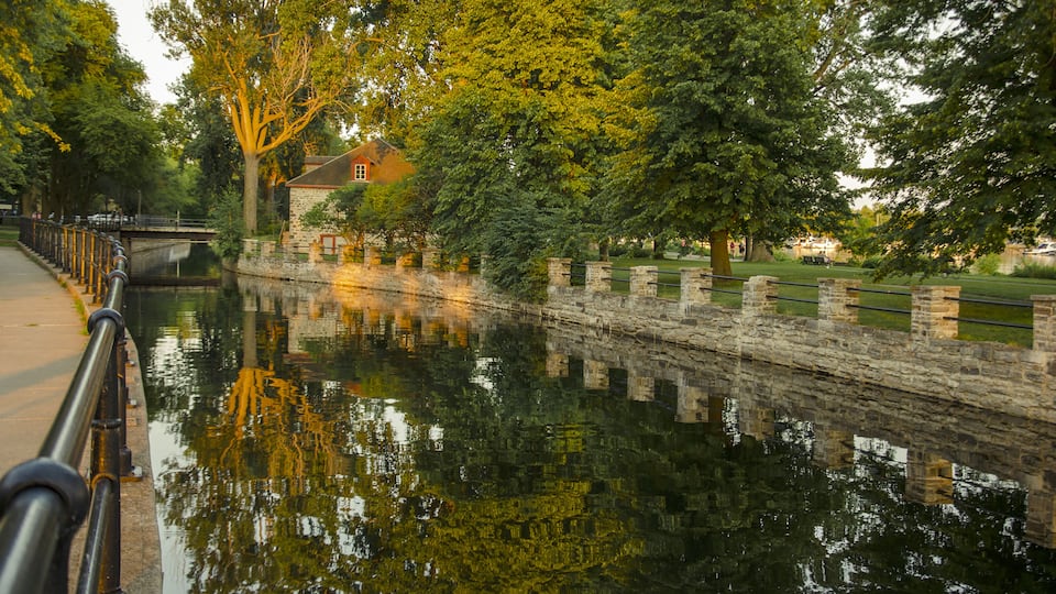 A canal with stone walls, trees, and a house in the background.