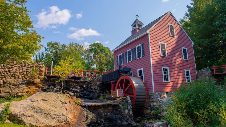 A red watermill with a wooden bridge, surrounded by greenery and a stone wall.