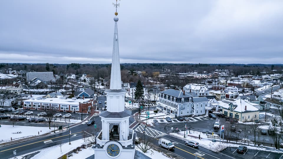 A town with a prominent white church steeple, a snow-covered landscape, and a mix of residential and commercial buildings.