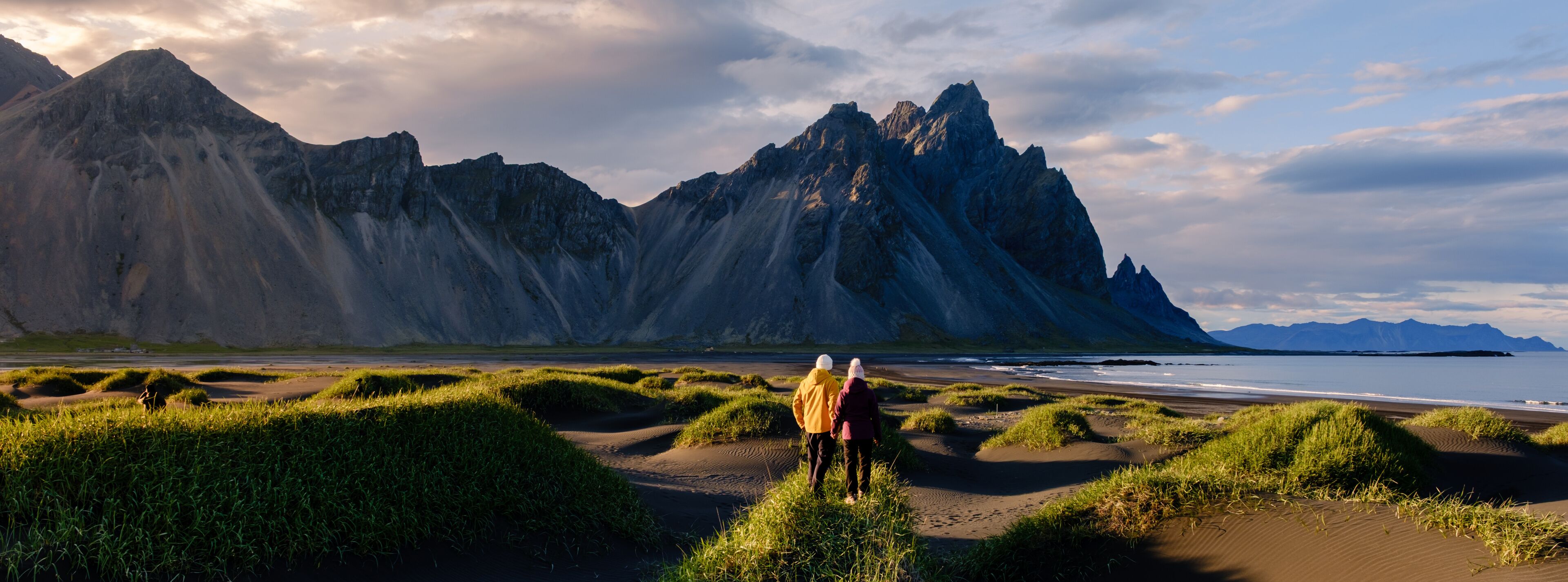 Two adventurers walk hand-in-hand on the striking black sand beach of Vestrahorn, surrounded by dramatic mountain peaks and vibrant green grass. Stokksnes beach Vestrahorn mountain Iceland