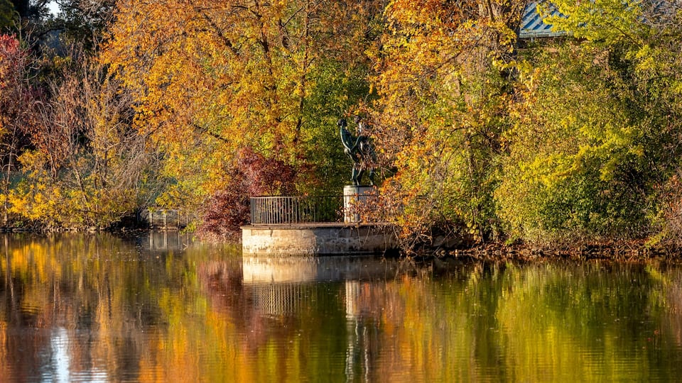 A serene lake with autumn foliage, a statue, and a bridge.
