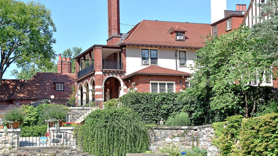 A large, historic house with a red-tiled roof, brick chimneys, and a stone staircase leading to a balcony.