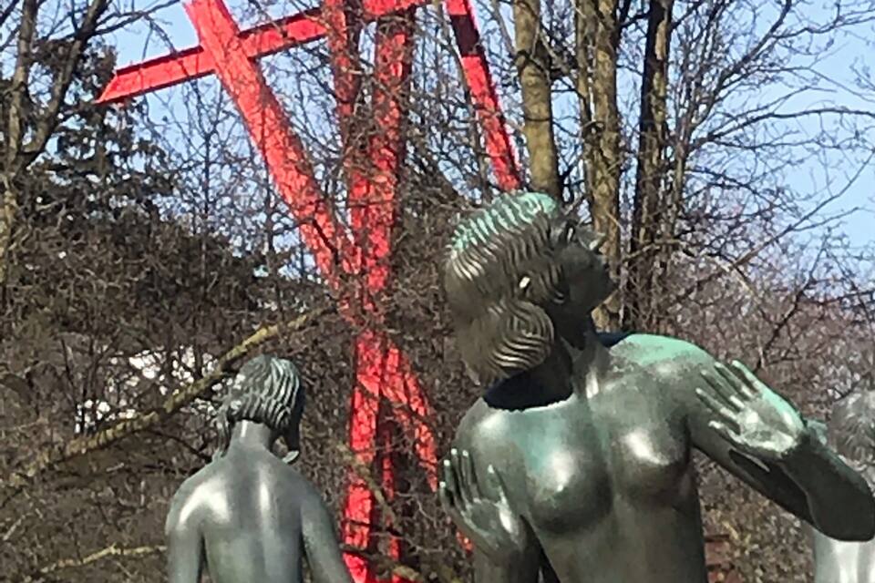 Escultura d'una dona amb un nen a un parc, amb una estructura vermella al fons.