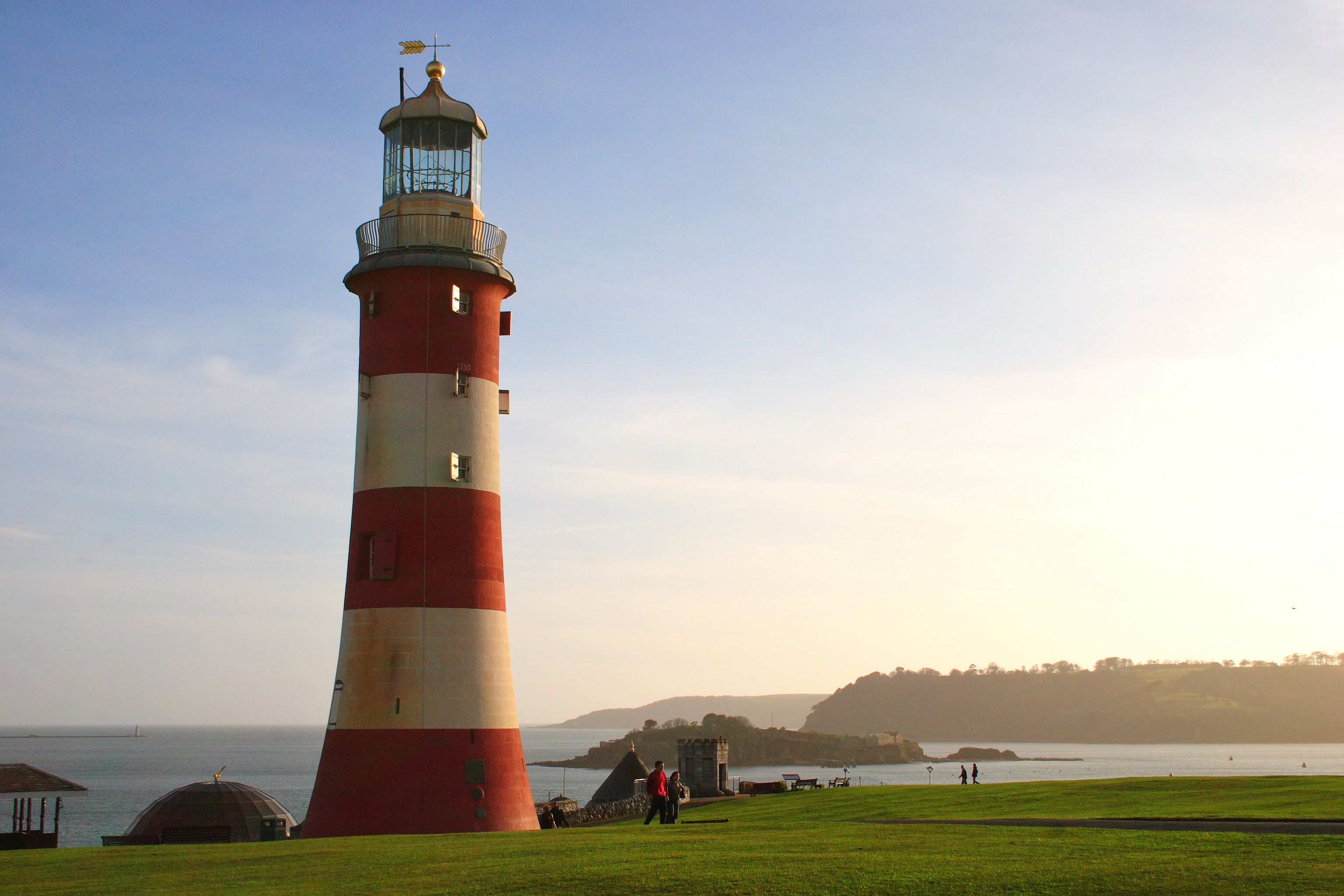 A red and white lighthouse with a weather vane on top, situated on a grassy hill overlooking the sea.