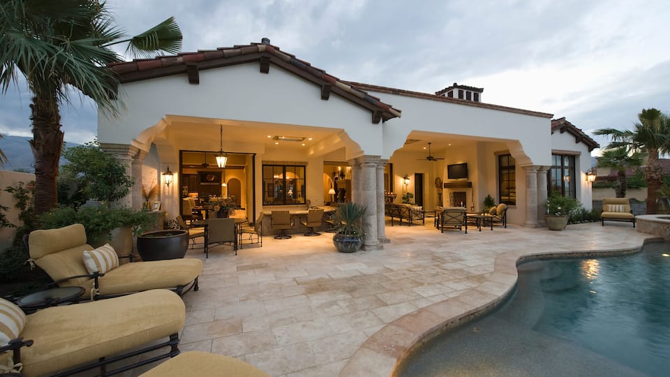 A Mediterranean-style house with a swimming pool, outdoor seating, and palm trees.