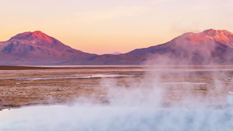 A geothermal area with steam vents and a lake surrounded by mountains.