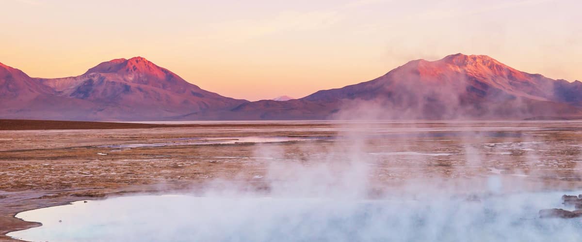 A geothermal area with steam vents and a lake surrounded by mountains.