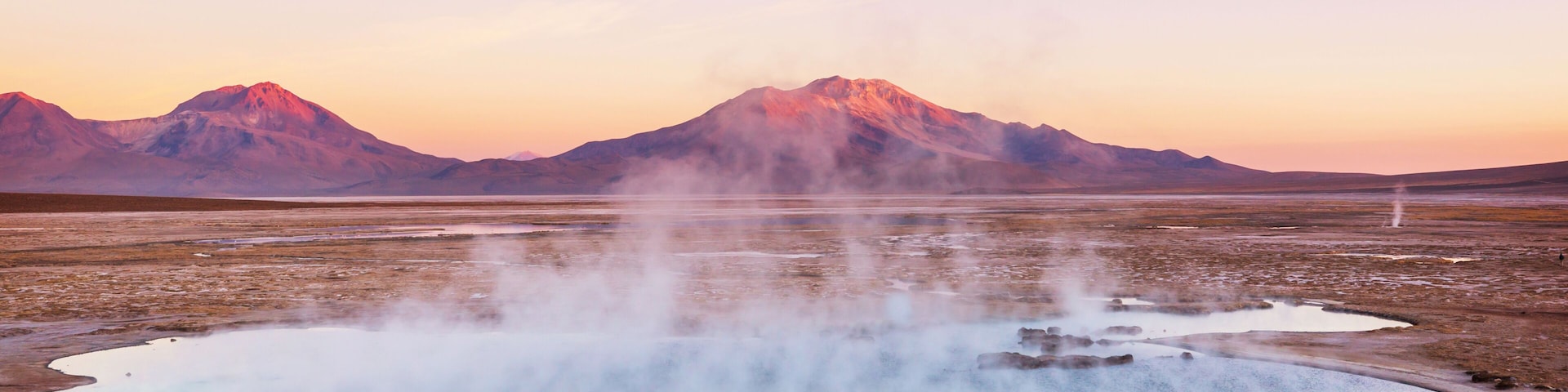 A geothermal area with steam vents and a lake surrounded by mountains.
