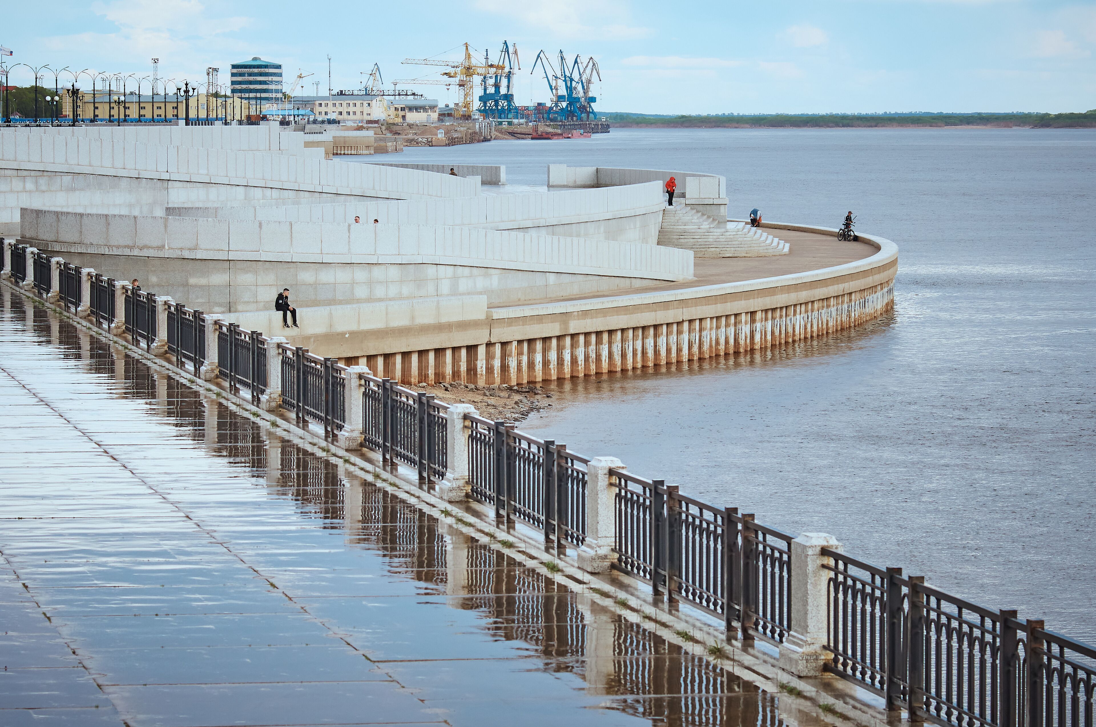 Wet river embankment after rain. Fencing, stairs and descents to the water. Cranes of the river port in the background. Blue cloudy sky. Citizens walk along the promenade. Reflections in puddles.