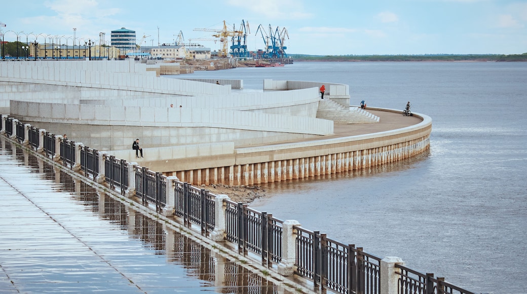 Wet river embankment after rain. Fencing, stairs and descents to the water. Cranes of the river port in the background. Blue cloudy sky. Citizens walk along the promenade. Reflections in puddles.
