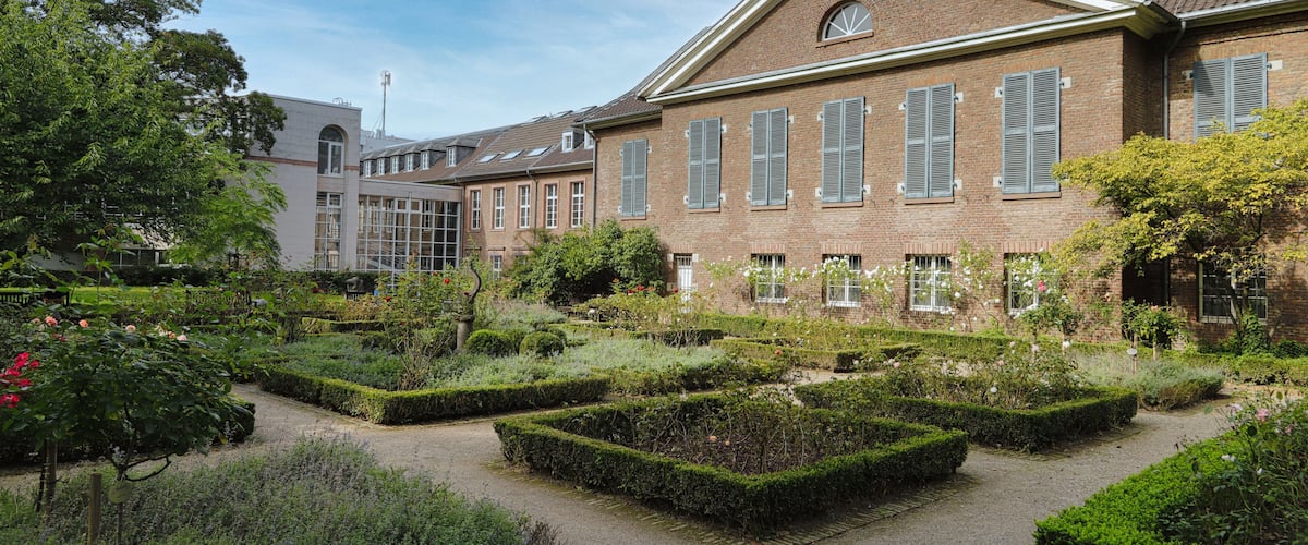 A courtyard with neatly trimmed hedges, a brick building with multiple windows, and a clear blue sky.
