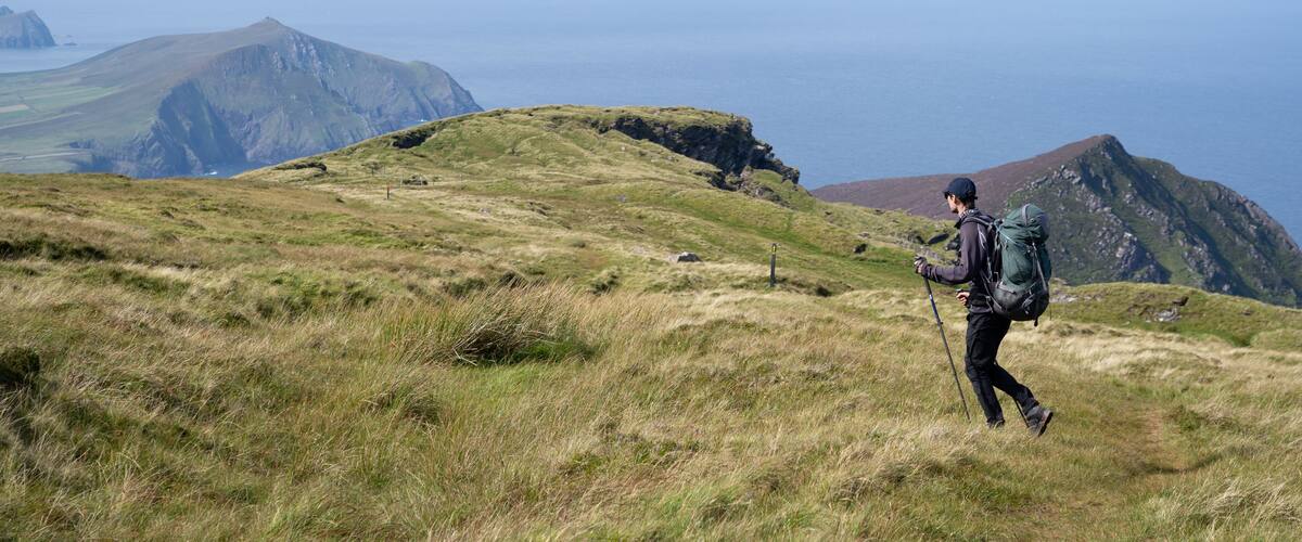 A hiker on a grassy hill with a mountainous backdrop and the ocean in the distance.