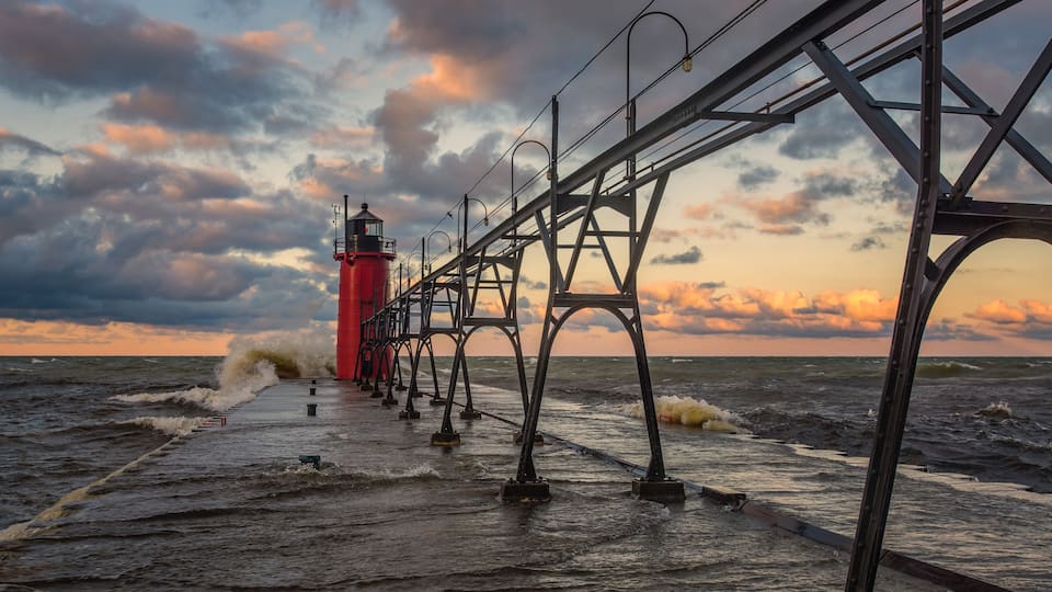 A red lighthouse on a pier with waves crashing against it.