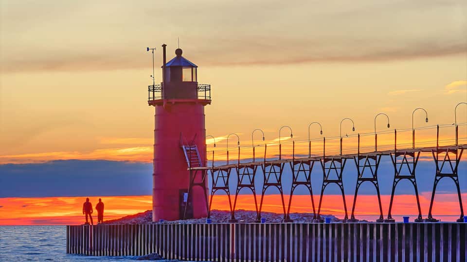 A lighthouse, a pier, and two people standing on the pier during sunset.