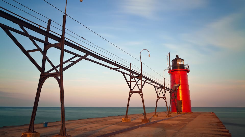 A red lighthouse on a pier with metal structures and a calm sea in the background.