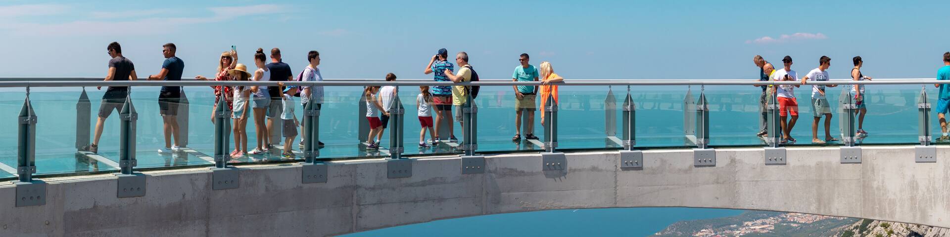 A glass bridge with people standing on it, overlooking a valley and a body of water.