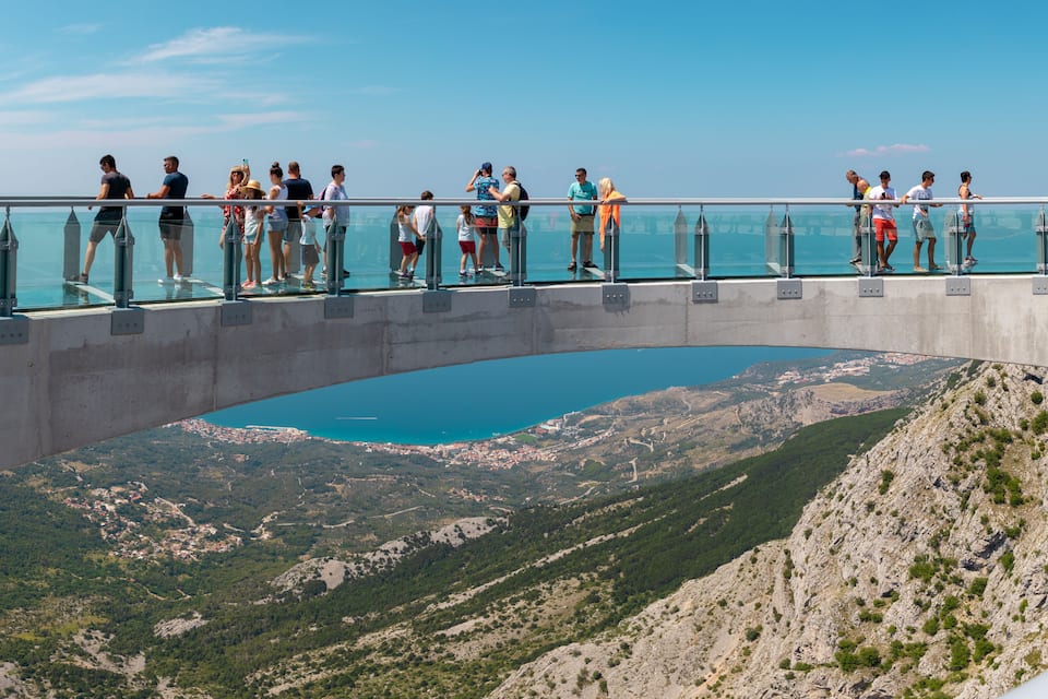 Un puente de cristal con gente, con vistas a un valle y un cuerpo de agua.