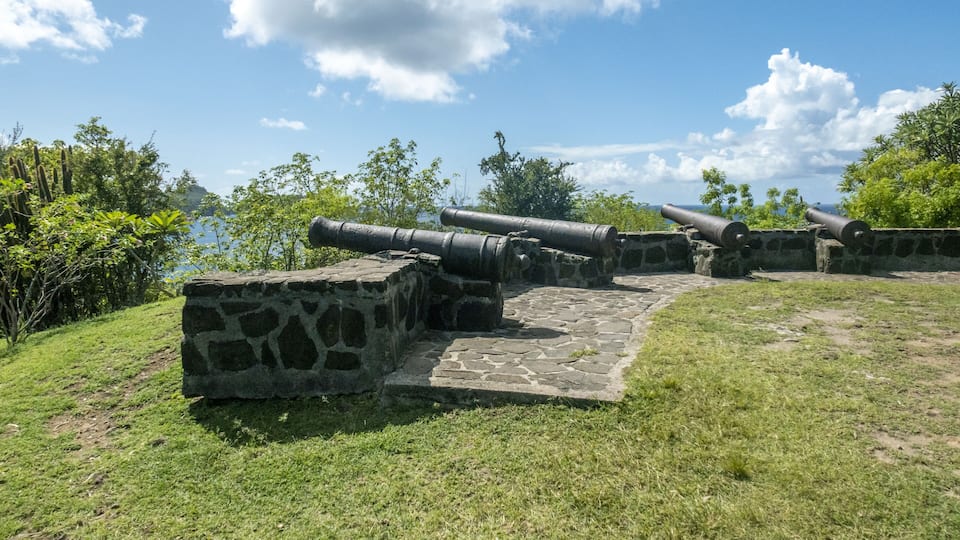 A stone cannon platform with three cannons overlooking a body of water, surrounded by greenery.