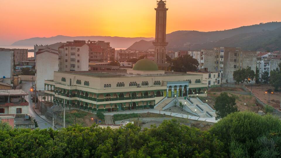 A historic building with a central dome and a tall tower, surrounded by modern buildings and a hilly landscape during sunset.