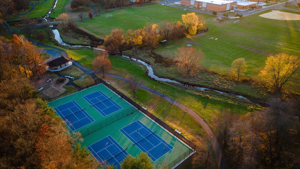 An aerial view of a sports complex with multiple tennis courts, a jogging path, and surrounding greenery.