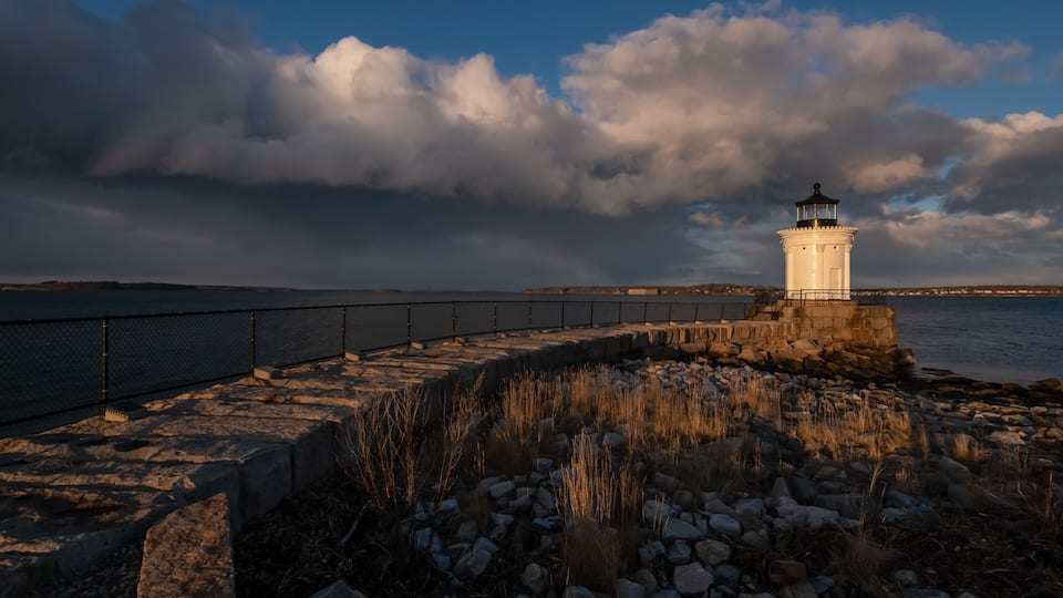 A lighthouse on a rocky outcrop by the sea, with a metal railing and a cloudy sky.