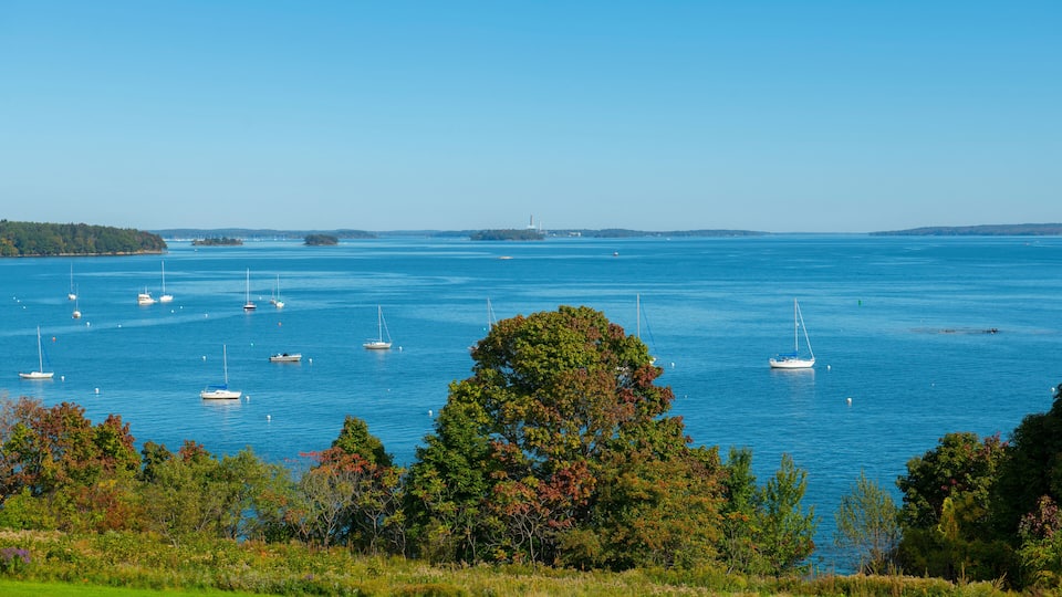 A coastal landscape with a clear blue sea, several sailboats, and a lighthouse in the distance.