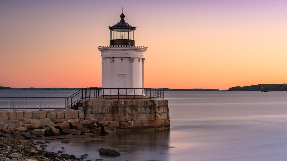 A white lighthouse on a rocky pier with a calm sea and a sunset sky.