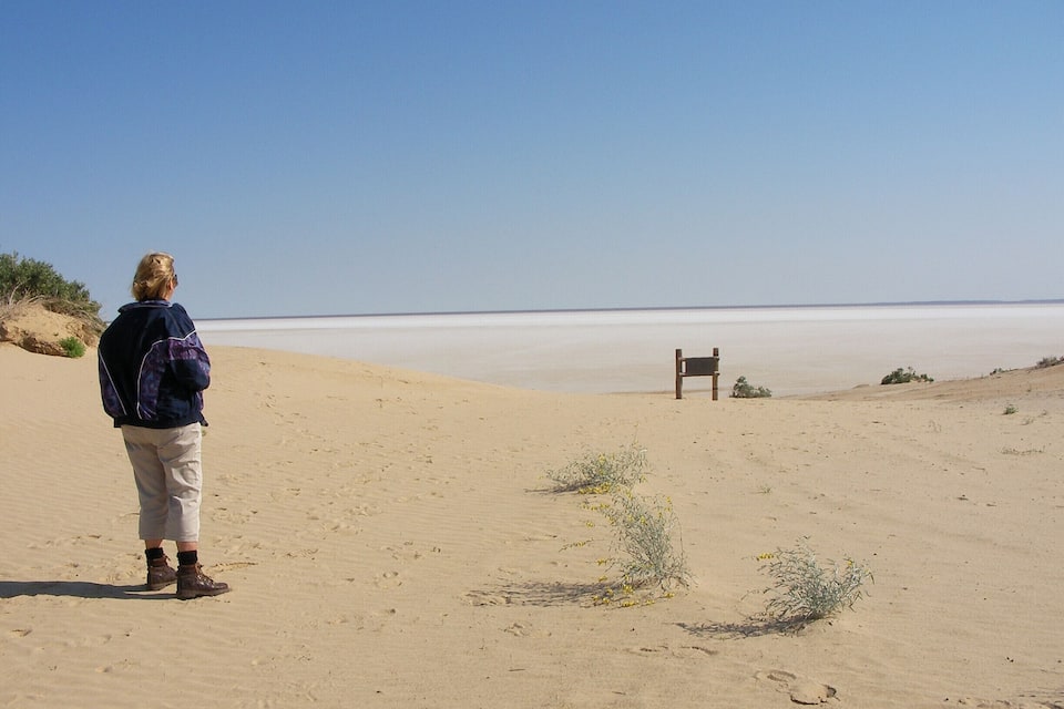 A person standing on a sandy beach with a bench in the distance.