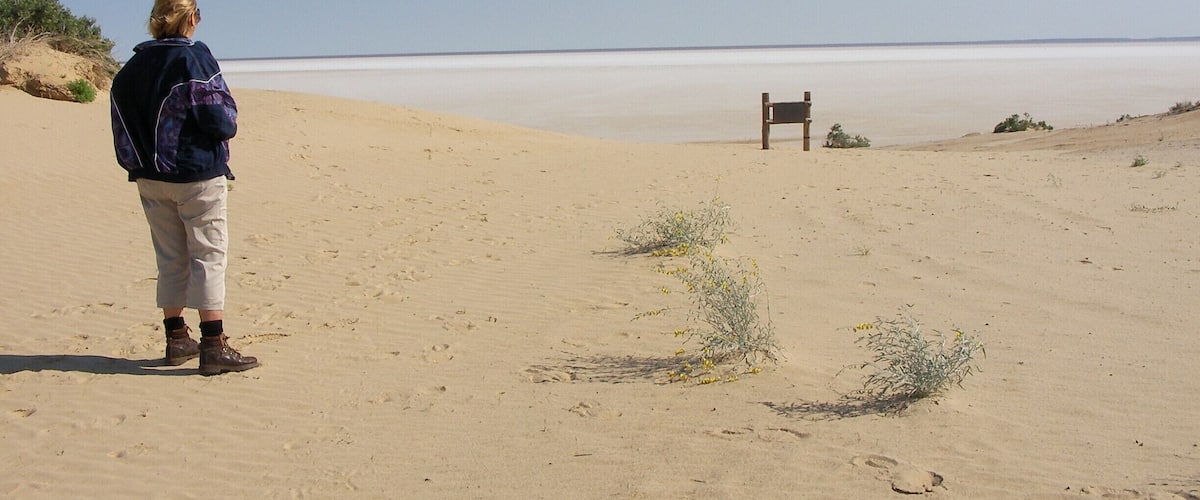 A person standing on a sandy beach with a bench in the distance.