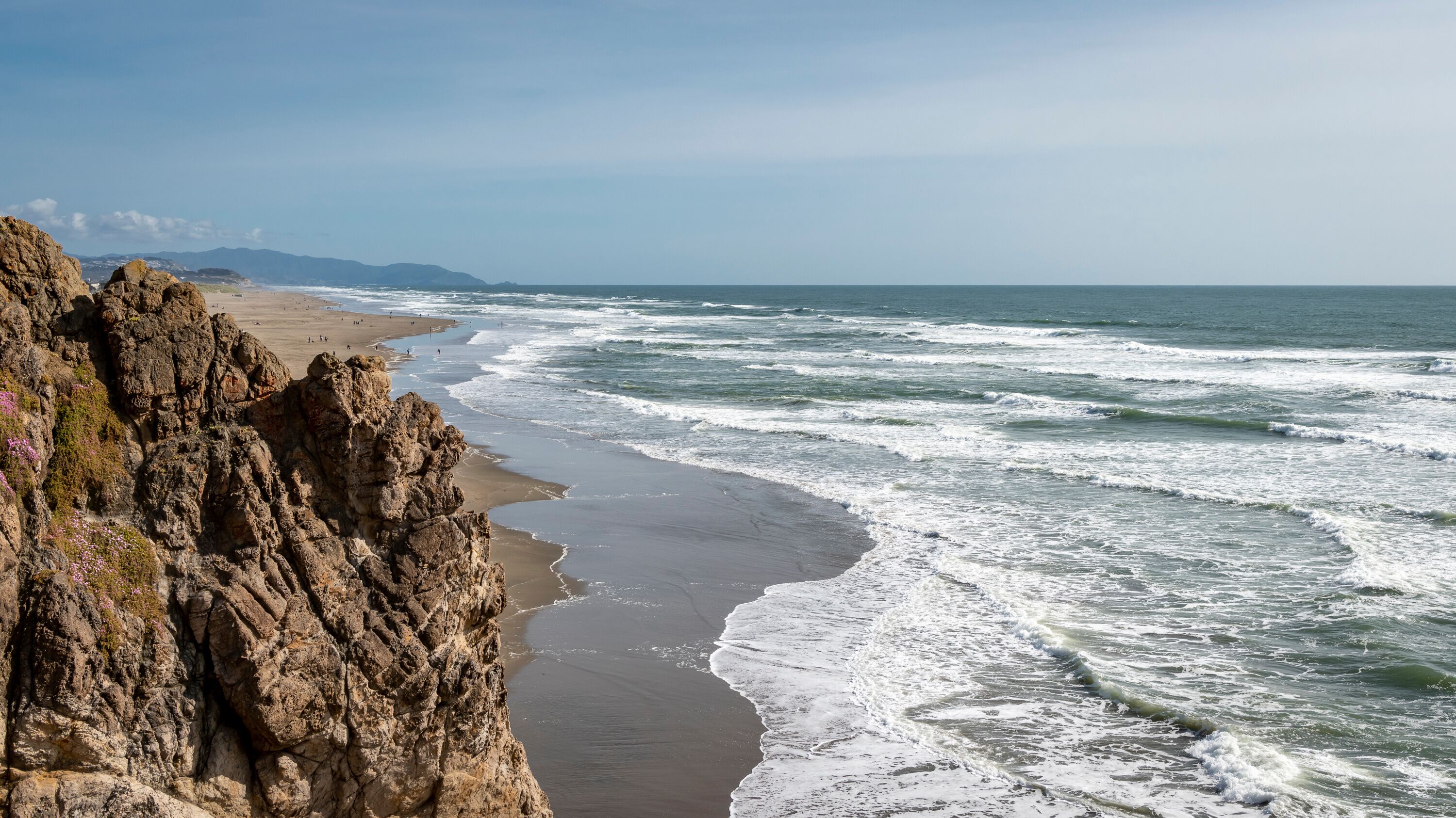 Spring afternoon at Ocean Beach in San Francisco.  High angle shot looking south from Cliff House.  Mild surf, people enjoying a relatively warm day.  Blue sky.  Rough cliffs foreground.