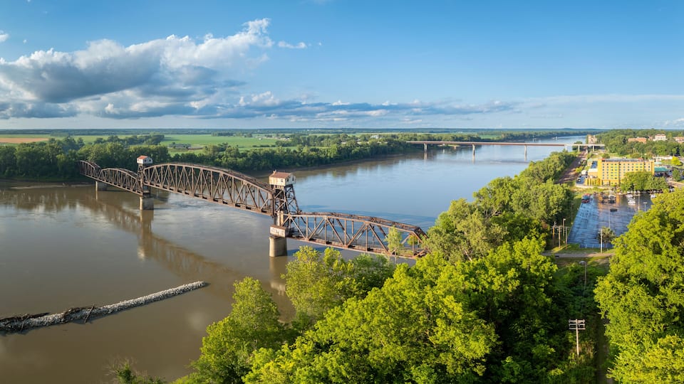 A large metal bridge over a river, surrounded by lush greenery and a distant cityscape.