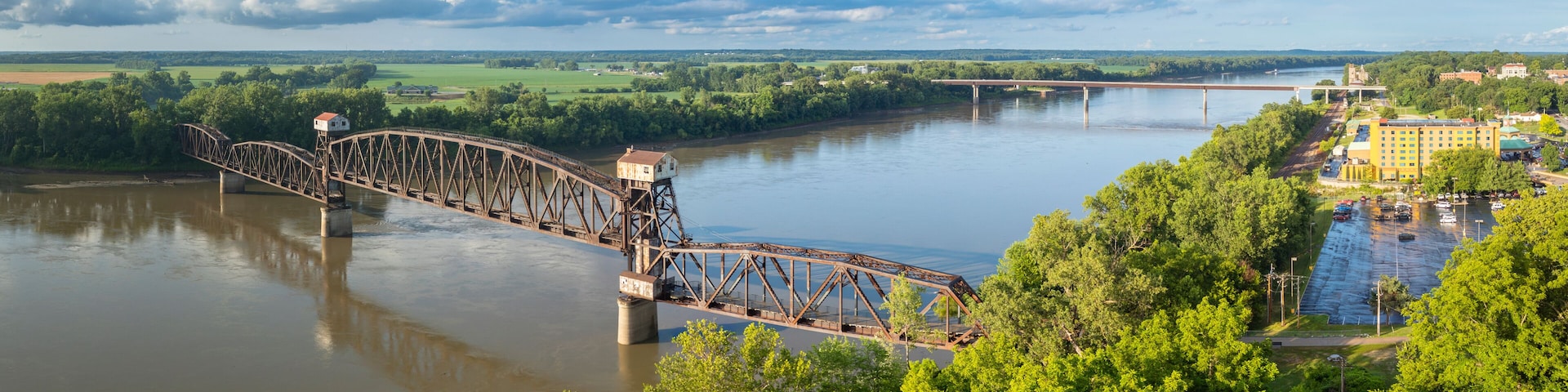 A large metal bridge over a river, surrounded by lush greenery and a distant cityscape.