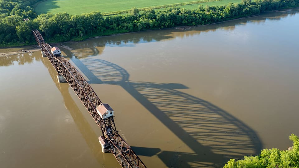 A large metal bridge spanning a wide river, surrounded by lush greenery and fields.
