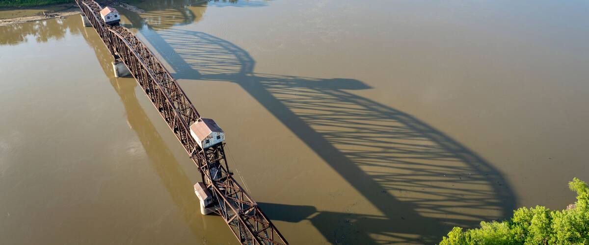 A large metal bridge spanning a wide river, surrounded by lush greenery and fields.