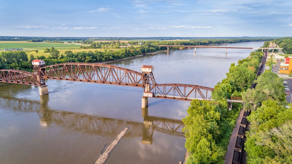 A large steel bridge spanning a wide river, surrounded by lush greenery and distant fields.