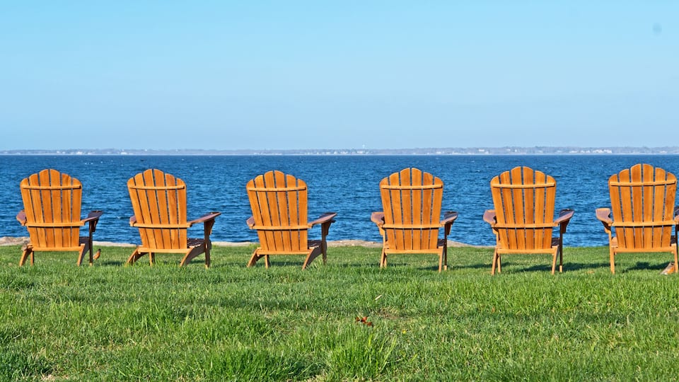 Six wooden Adirondack chairs lined up on a grassy field by a body of water.