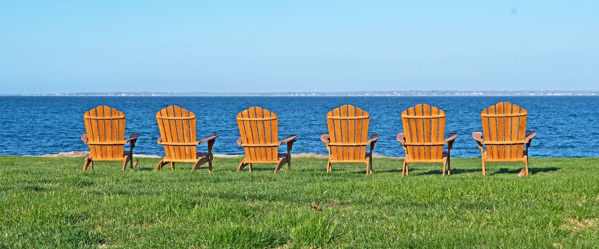 Six wooden Adirondack chairs lined up on a grassy field by a body of water.
