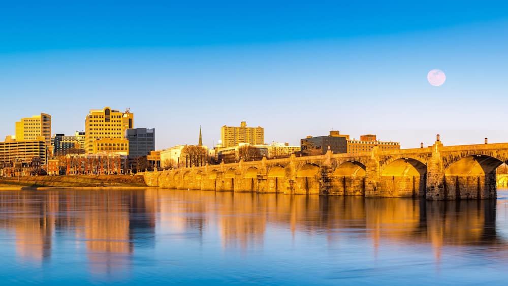 A city skyline with a historic stone bridge over a calm river, reflecting the buildings and the moon in the sky.