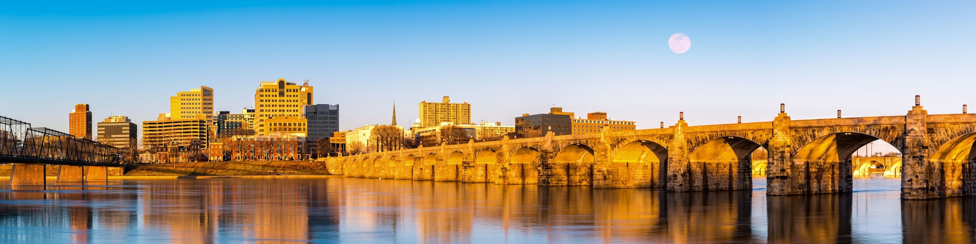 A city skyline with a historic stone bridge over a calm river, reflecting the buildings and the moon in the sky.